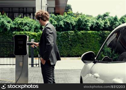 Progressive businessman with electric car recharging at public charging station at modern city residential area. Eco friendly rechargeable car powered by alternative clean energy.. Progressive businessman charging EV car at public parking car charging station.