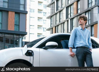 Progressive businessman with electric car recharging at public parking car charging station at modern city center. Eco friendly rechargeable car powered by alternative clean energy.. Progressive businessman with EV car at public parking car charging station.