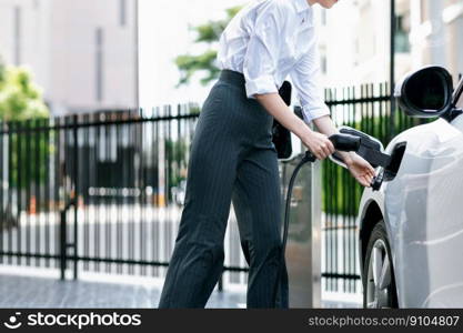 Progressive businesswoman insert charger plug from charging station to her electric vehicle with apartment condo building in background. Eco friendly rechargeable car powered by sustainable energy.. Progressive businesswoman plugs charger plug from charging station to EV.