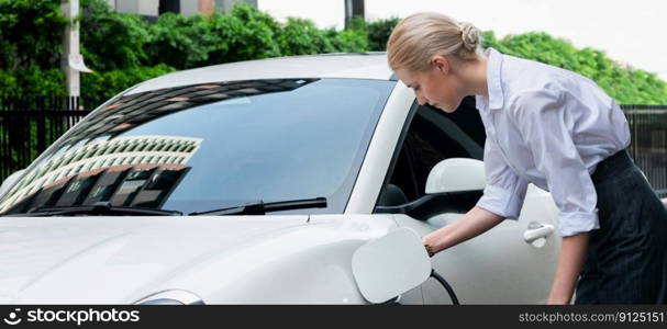 Progressive businesswoman insert charger plug from charging station to her electric vehicle with apartment condo building in background. Eco friendly rechargeable car powered by sustainable energy.. Progressive businesswoman plugs charger plug from charging station to EV.