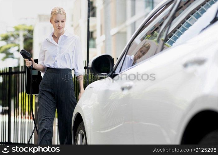Progressive businesswoman insert charger plug from charging station to her electric vehicle with apartment condo building in background. Eco friendly rechargeable car powered by sustainable energy.. Progressive businesswoman plugs charger plug from charging station to EV.