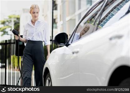 Progressive businesswoman insert charger plug from charging station to her electric vehicle with apartment condo building in background. Eco friendly rechargeable car powered by sustainable energy.. Progressive businesswoman plugs charger plug from charging station to EV.