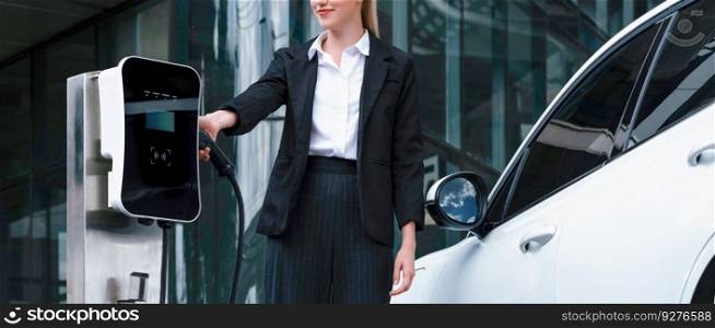 Progressive businesswoman wearing suit with electric car recharging at public parking car charging station at modern city center. Eco friendly rechargeable car powered by alternative clean energy.. Progressive businesswoman with EV car at public parking car charging station.