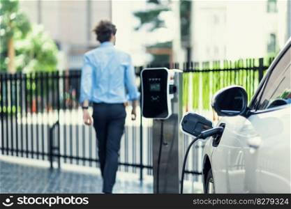 Progressive eco-friendly concept of focus parking EV car at public electric-powered charging station in city center with blur businessman walking in the background.. Progressive concept of focus EV car at charging station with blur man background