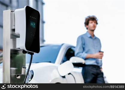 Progressive eco-friendly concept of focus parking EV car at public electric-powered charging station in city with blur background of businessman leaning on recharging-electric vehicle with coffee.. Progressive concept of focus EV car at charging station with blur man background