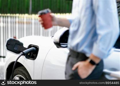 Progressive eco-friendly concept of focus parking EV car at public electric-powered charging station in city with blur background of businessman waiting on recharging car with coffee.. Focus EV car charging batter with progressive image of business man and coffee.