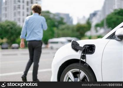 Progressive eco-friendly concept of focus parking EV car at public electric-powered charging station in city with blur background of businessman talking on the phone while recharging electric vehicle.. Progressive concept of focus EV car at charging station with blur man background