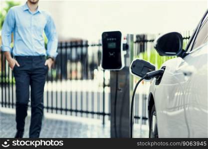 Progressive eco-friendly concept of focus parking EV car at public electric-powered charging station in city center with blur background of businessman waiting at his recharging-electric vehicle.. Progressive concept of focus EV car at charging station with blur man background