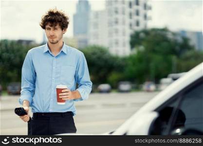 Progressive eco-friendly concept of parking EV car at public electric-powered charging station in city with blur background of businessman leaning on recharging-electric vehicle with coffee.. Progressive concept of EV car at charging station with blur man background