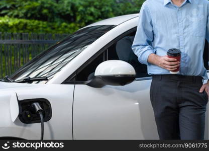 Progressive eco-friendly concept of parking EV car at public electric-powered charging station in city with blur background of businessman leaning on recharging-electric vehicle with coffee.. Progressive businessman with coffee and EV car at charging station.