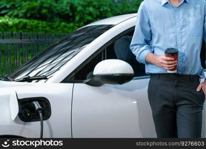 Progressive eco-friendly concept of parking EV car at public electric-powered charging station in city with blur background of businessman leaning on recharging-electric vehicle with coffee.. Progressive businessman with coffee and EV car at charging station.