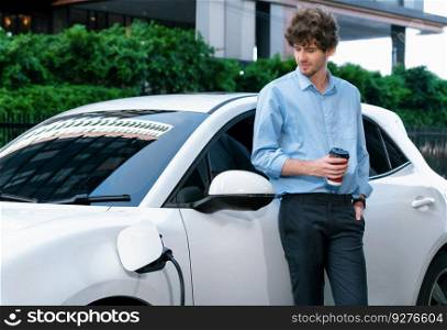 Progressive eco-friendly concept of parking EV car at public electric-powered charging station in city with blur background of businessman leaning on recharging-electric vehicle with coffee.. Progressive concept of EV car at charging station with blur man background