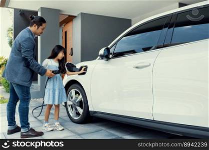 Progressive father and daughter plugs EV charger from home charging station to electric vehicle. Future eco-friendly car with EV cars powered by renewable source of clean energy.. Progressive dad and daughter charging EV car from home charging station.