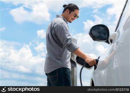 Progressive man with e≤ctric vehic≤and cloudscape background. EV car driven by c≤an re≠wab≤plug≥d-in with charging po∫. Refreshing environment from green e≠rgy.. Progressive man with plug≥d-in EV car with charging po∫, cloudscape backdrop.