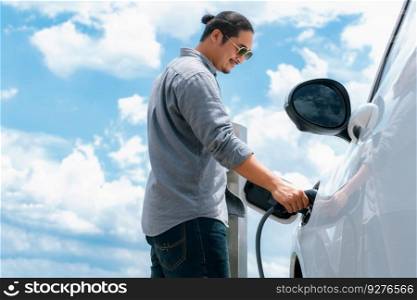 Progressive man with electric vehicle and cloudscape background. EV car driven by clean renewable plugged-in with charging point. Refreshing environment from green energy.. Progressive man with plugged-in EV car with charging point, cloudscape backdrop.