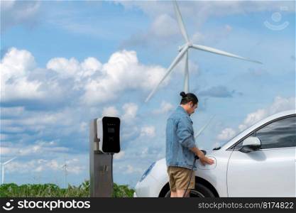 Progressive man with his electric car, EV car recharging energy from charging station on green field with wind turbine as concept of future sustainable energy. Electric vehicle with energy generator.. Progressive man with his EV car and wind turbine as concept of renewable energy.