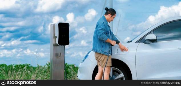 Progressive man with his electric car, EV car recharging energy from charging station on green field with wind turbine as concept of future sustainable energy. Electric vehicle with energy generator.. Progressive man with his EV car and wind turbine as concept of renewable energy.