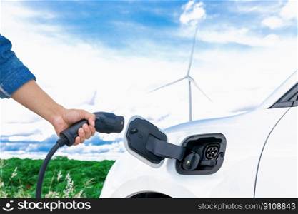Progressive natural scenic with windmill generator where hand insert charging plug to electric vehicle from charging station with natural background. EV car powered by wind turbine electric generator.. Hand insert connector plug to EV with progressive natural scenic and windturbine