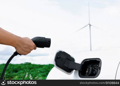 Progressive natural scenic with windmill generator where hand insert charging plug to electric vehicle from charging station with natural background. EV car powered by wind turbine electric generator.. Hand insert connector plug to EV with progressive natural scenic and windturbine