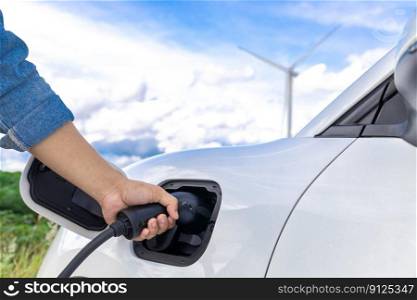 Progressive natural scenic with windmill generator where hand insert charging plug to electric vehicle from charging station with natural background. EV car powered by wind turbine electric generator.. Hand insert connector plug to EV with progressive natural scenic and windturbine