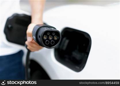 Progressive new alternative clean energy vehicle and electric charging device concept. Focus closeup hand holding and pointing EV charger plug at camear from electric charging station at camera. Progressive idea of focus hand pointing charging plug at camera, blur background