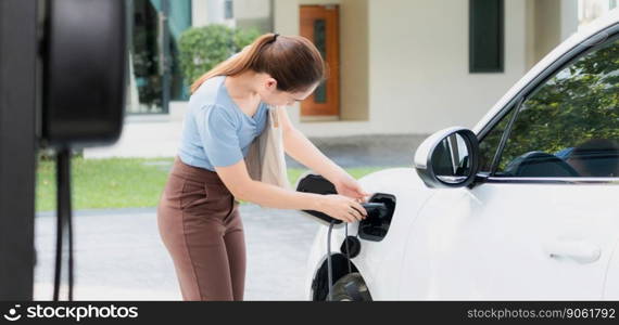 Progressive woman install cable plug to her electric car with home charging station. Concept of the use of electric vehicles in a progressive lifestyle contributes to clean environment.. Progressive woman recharge her EV car at home charging station.