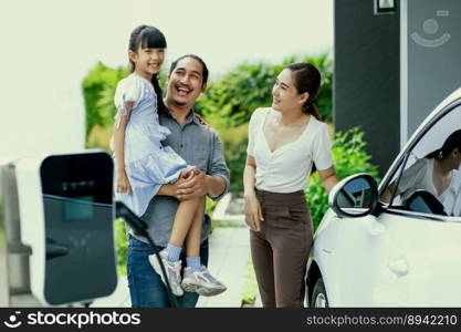 Progressive young parent teach daughter how to recharge or refuel EV car at home charging station. Green and clean energy from electric vehicle for healthy environment. Eco power from renewable source. Progressive young parents and daughter living in a home with an electric car.