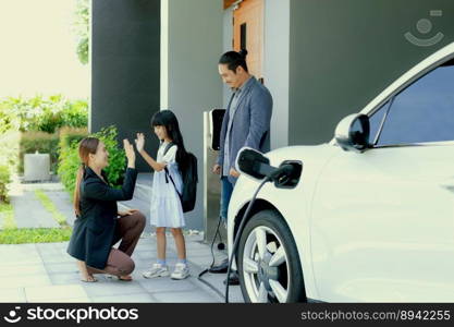 Progressive young parents and daughter with electric vehicle and home charging station. Green and clean energy from electric vehicles for healthy environment. Eco power from renewable source at home.. Progressive young parents and daughter living in a home with an electric car.