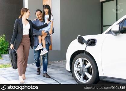 Progressive young parents and daughter with electric vehicle and home charging station. Green and clean energy from electric vehicles for healthy environment. Eco power from renewable source at home.. Progressive young parents and daughter living in a home with an electric car.