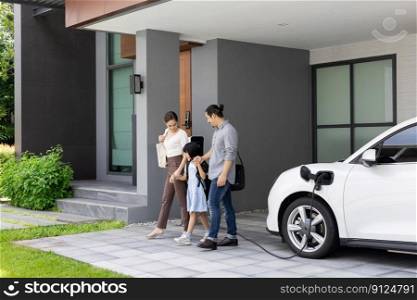 Progressive young parents and daughter with electric vehicle and home charging station. Green and clean energy from electric vehicles for healthy environment. Eco power from renewable source at home.. Progressive young parents and daughter living in a home with an electric car.