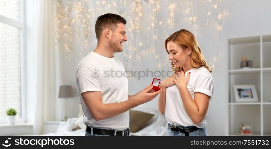 proposal, christmas and valentine&rsquo;s day concept - man giving diamond engagement ring in little red box to happy woman over bedroom with festive garland lights on background. man giving woman engagement ring on valentines day