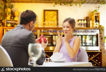 proposal, couple and valentine&rsquo;s day concept - excited young woman looking at man with engagement ring over restaurant or wine bar background. man making proposal to happy woman at restaurant