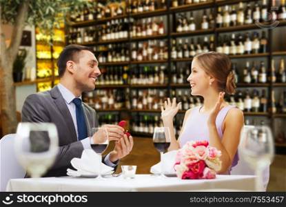 proposal, couple and valentine&rsquo;s day concept - excited young woman looking at man with engagement ring over restaurant or wine bar background. man making proposal to happy woman at restaurant
