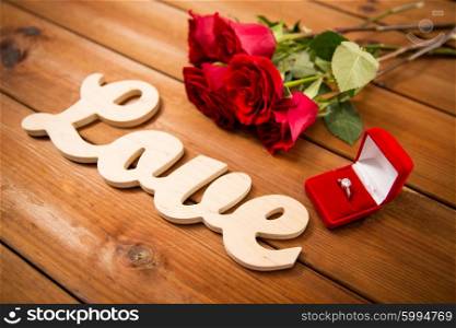 proposal, romance, valentines day and holidays concept - close up of gift box with diamond engagement ring, red roses and word love on wood