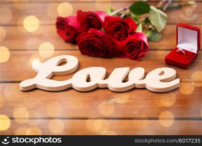 proposal, romance, valentines day and holidays concept - close up of gift box with diamond engagement ring, red roses and word love on wood over golden lights