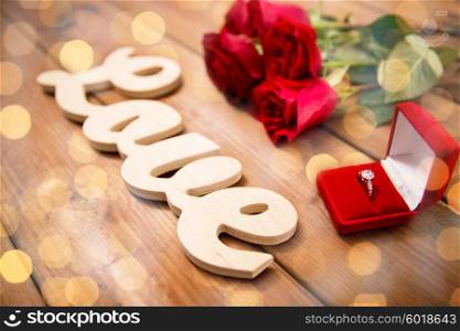 proposal, romance, valentines day and holidays concept - close up of gift box with diamond engagement ring, red roses and word love on wood