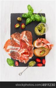prosciutto ham with bread, basil pesto and tomatoes on slate and white wooden background, top view