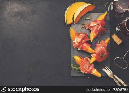 Prosciutto ham with cantaloupe melon, basil leaves and wine over dark grunge background. Top view, copy space