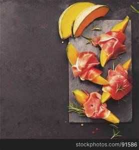 Prosciutto ham with cantaloupe melon, basil leaves and wine over dark grunge background. Top view, copy space