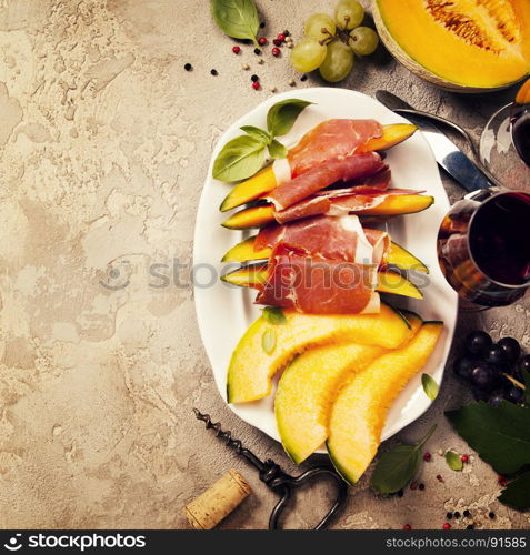 Prosciutto ham with cantaloupe melon, basil leaves and wine over grunge background. Top view, copy space