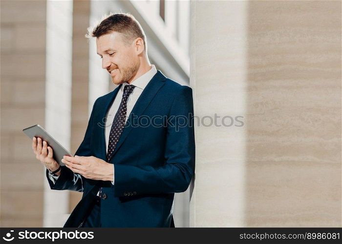 Prosperous businessman in formal clothes reads notification on digital tablet, installs new application, connected to wireless internet, poses indoor. People, business and modern technology concept