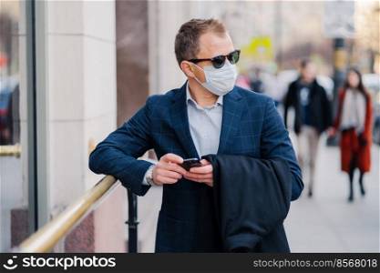 Prosperous businessman in formal wear poses at street, waits for someone, holds mobile phone and sends text messages, wears medical mask during coronavirus outbreak, few people walking outside