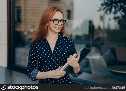 Prosperous businesswoman with red hair carries modern device and notepad goes on formal meeting wears spectacles black polka dot dress looks happily into distance poses outdoor near restaurant. Businesswoman with red hair carries modern device and notepad goes on formal meeting