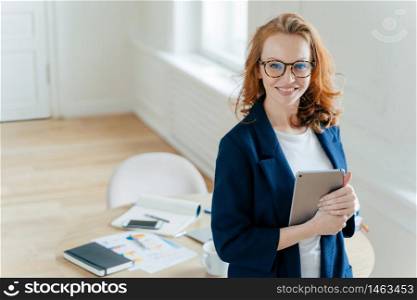 Prosperous female leader of working team holds digital tablet device, develops business ideas, has toothy smile, red hair, wears elegant outfit, stands in own cabinet, involved in working process