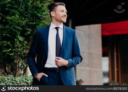 Prosperous handsome businessman in formal suit, keeps hand in pocket, looks positively aside, stands near bank, has cheerful expression, positive smile. People, business and finances concept.