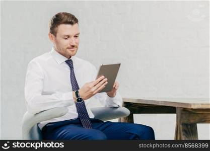 Prosperous handsome young businessman watches webinar online on modern tablet computer, dressed in formal clothes, sits at chair indoor. Young attractive male financier reads news on web page online