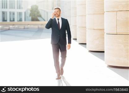 Prosperous male business owner in formal suit, has telephone conversation with business partner while walks to meeting in urban setting, satisfied to hear good news. People and career concept