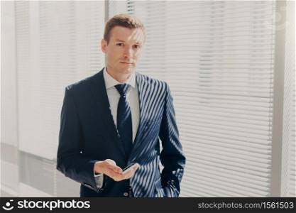 Prosperous male director sends message and multimedia files via smart phone, synchronizes phone data and shares photos for project, poses in office, wears luxury expensive suit, looks seriously