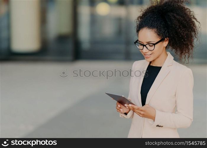 Prosperous owner of business company stands with digital touchpad, focused in screen, searches for new ideas of developing market, wears glasses and white formal jacket. Businesswoman outdoor