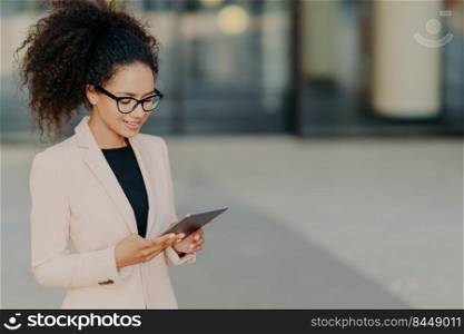 Prosperous owner of business company stands with digital touchpad, focused in screen, searches for new ideas of developing market, wears glasses and white formal jacket. Businesswoman outdoor